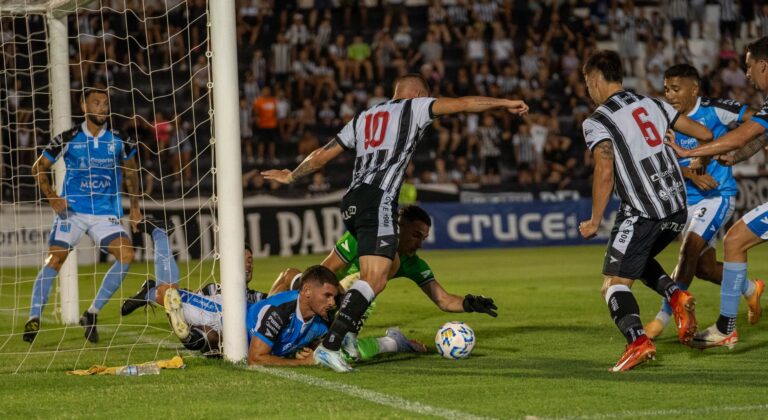 Dramatic goal-line action during a night soccer match, players in black and blue jerseys.