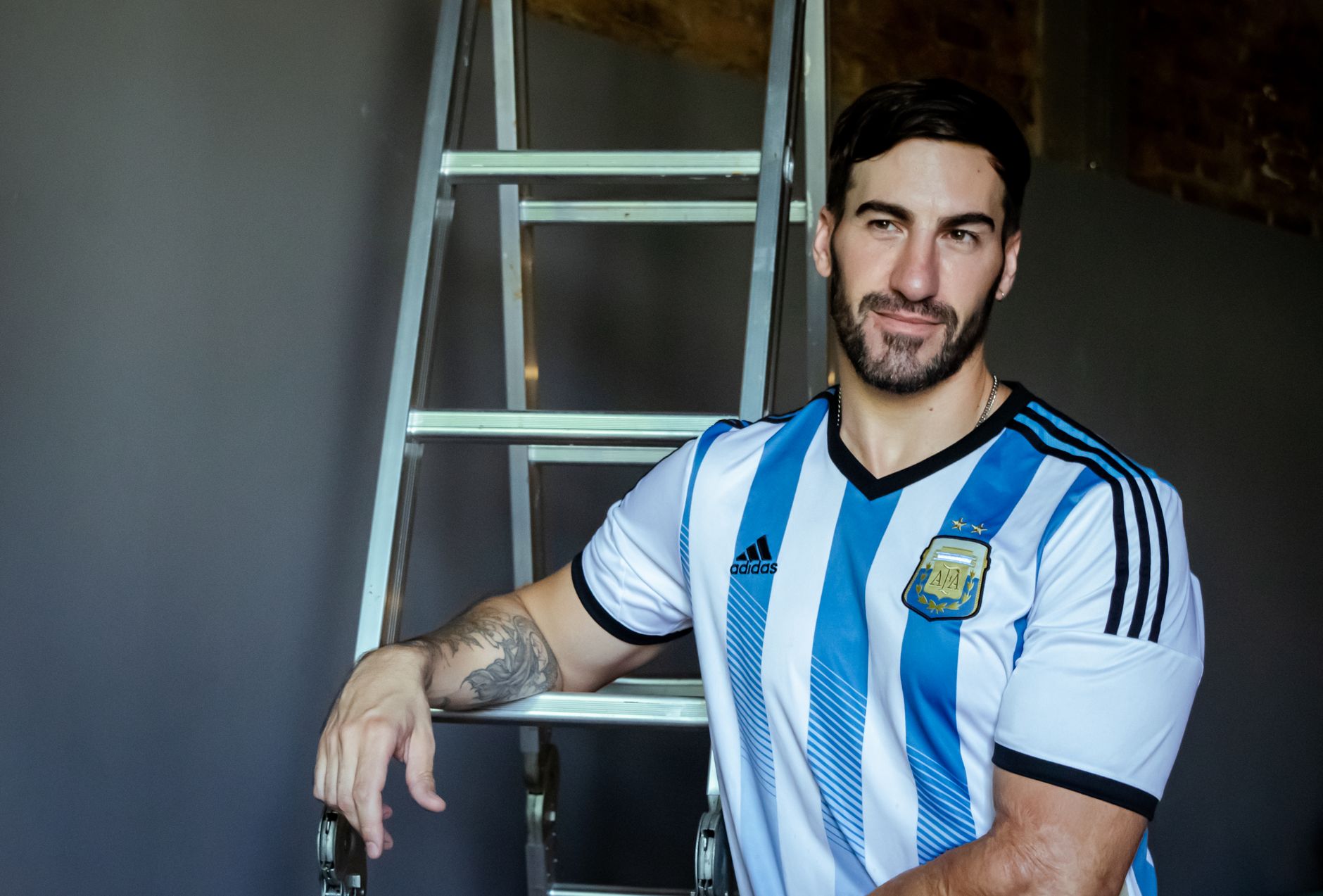 Portrait of a man in an Argentina football jersey standing beside a ladder indoors.
