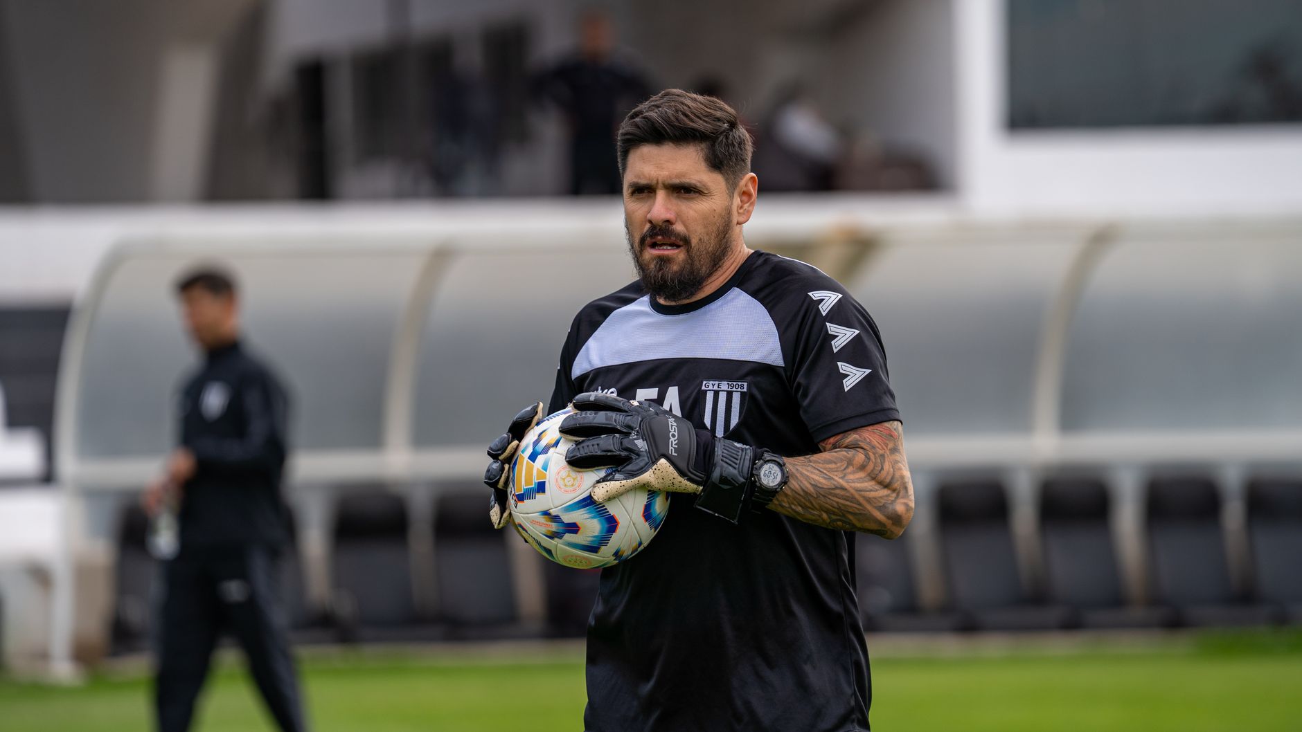 A focused football coach holding a soccer ball on the field during daytime practice.