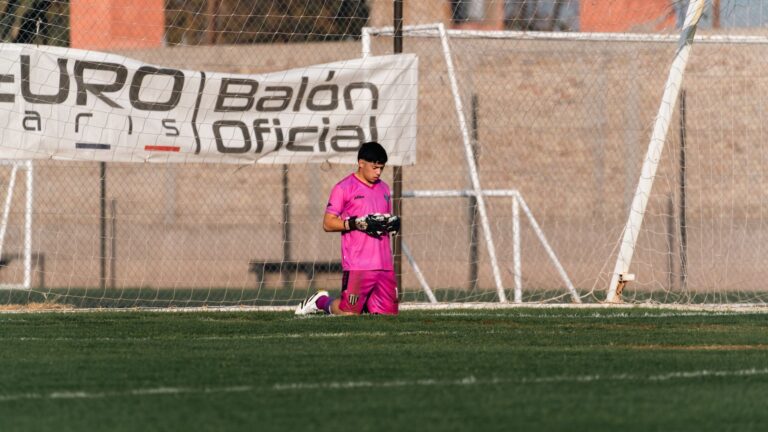 Goalkeeper kneeling on field in pink kit during a soccer match outdoors.