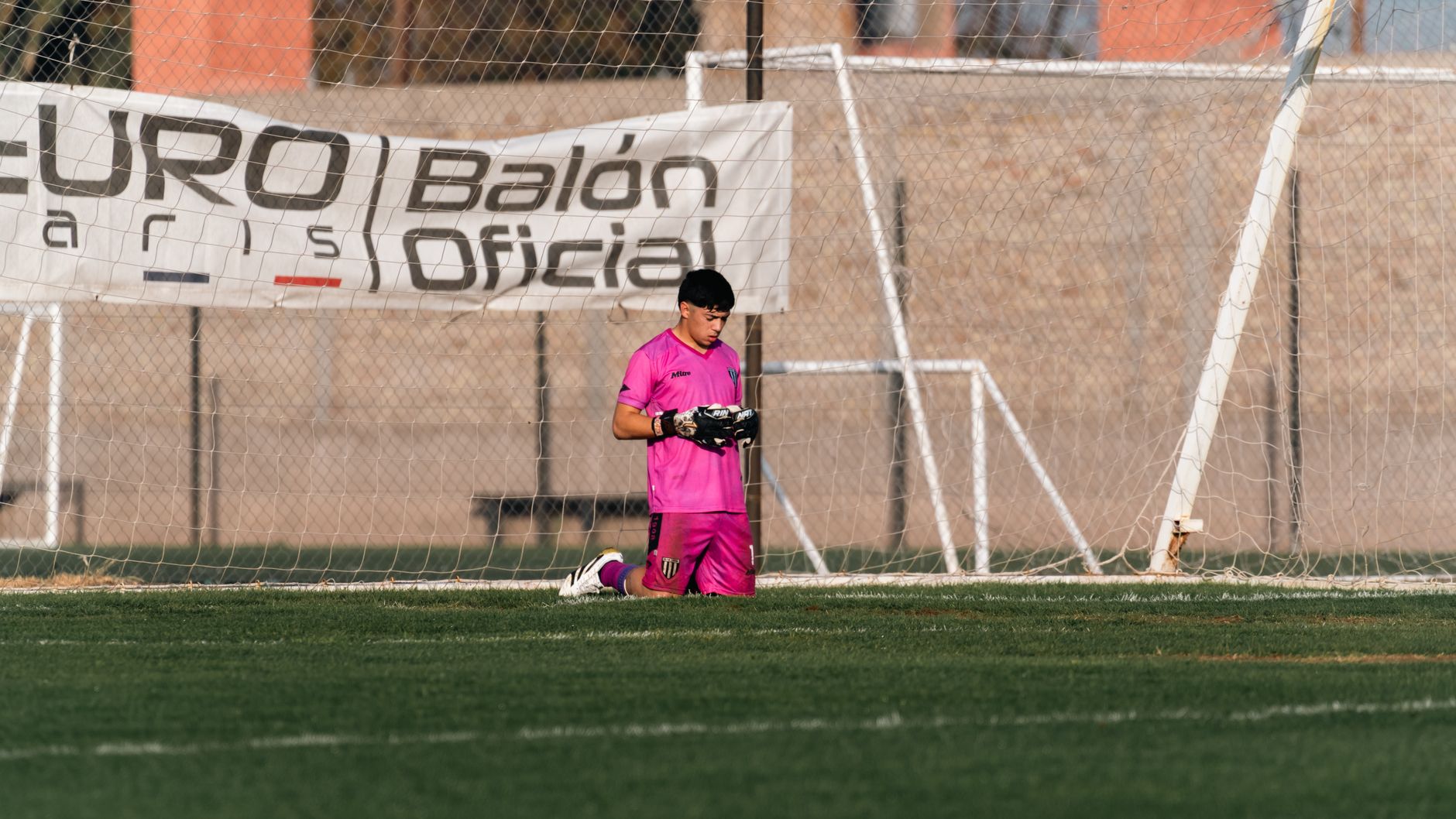Goalkeeper kneeling on field in pink kit during a soccer match outdoors.
