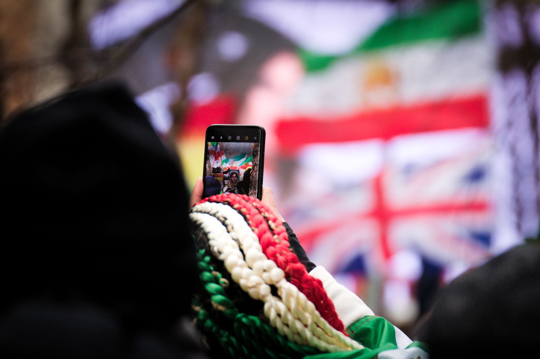 Person using smartphone to capture live event in Brussels with visible flags in background.