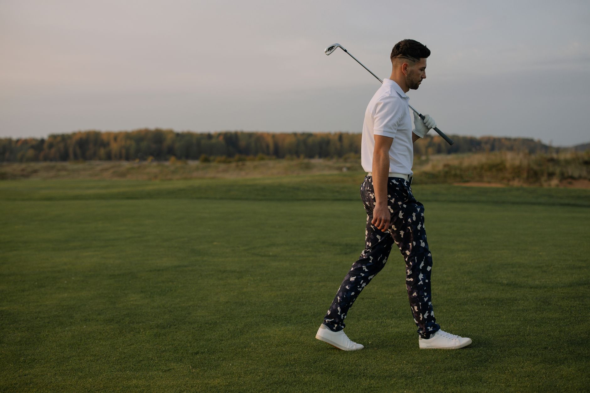 A stylish male golfer walks on the golf course during sunset carrying a golf club.