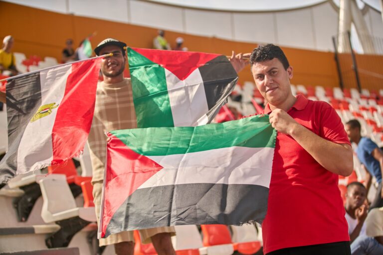 Enthusiastic soccer fans holding Egyptian and Palestinian flags at a sports stadium.