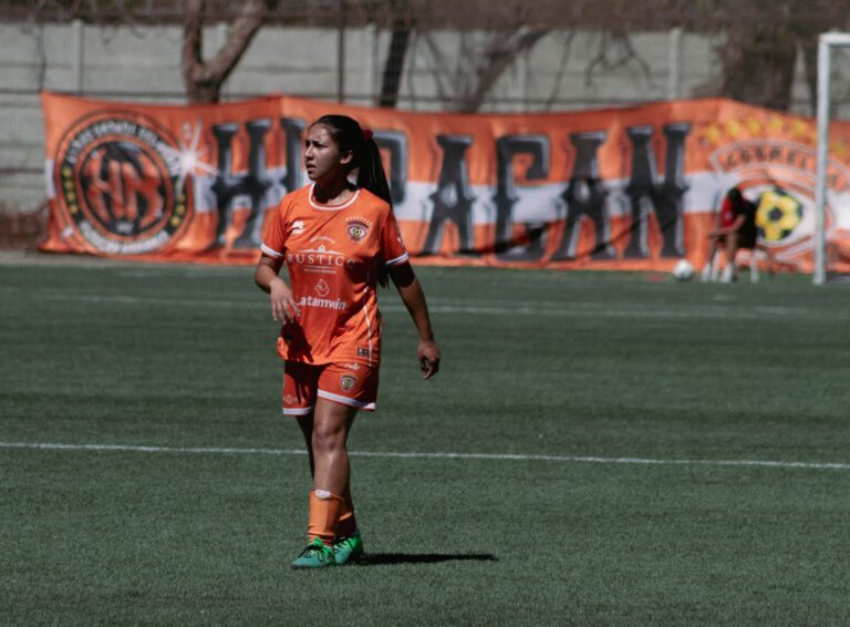 Female soccer player in orange uniform on outdoor field with banner background.
