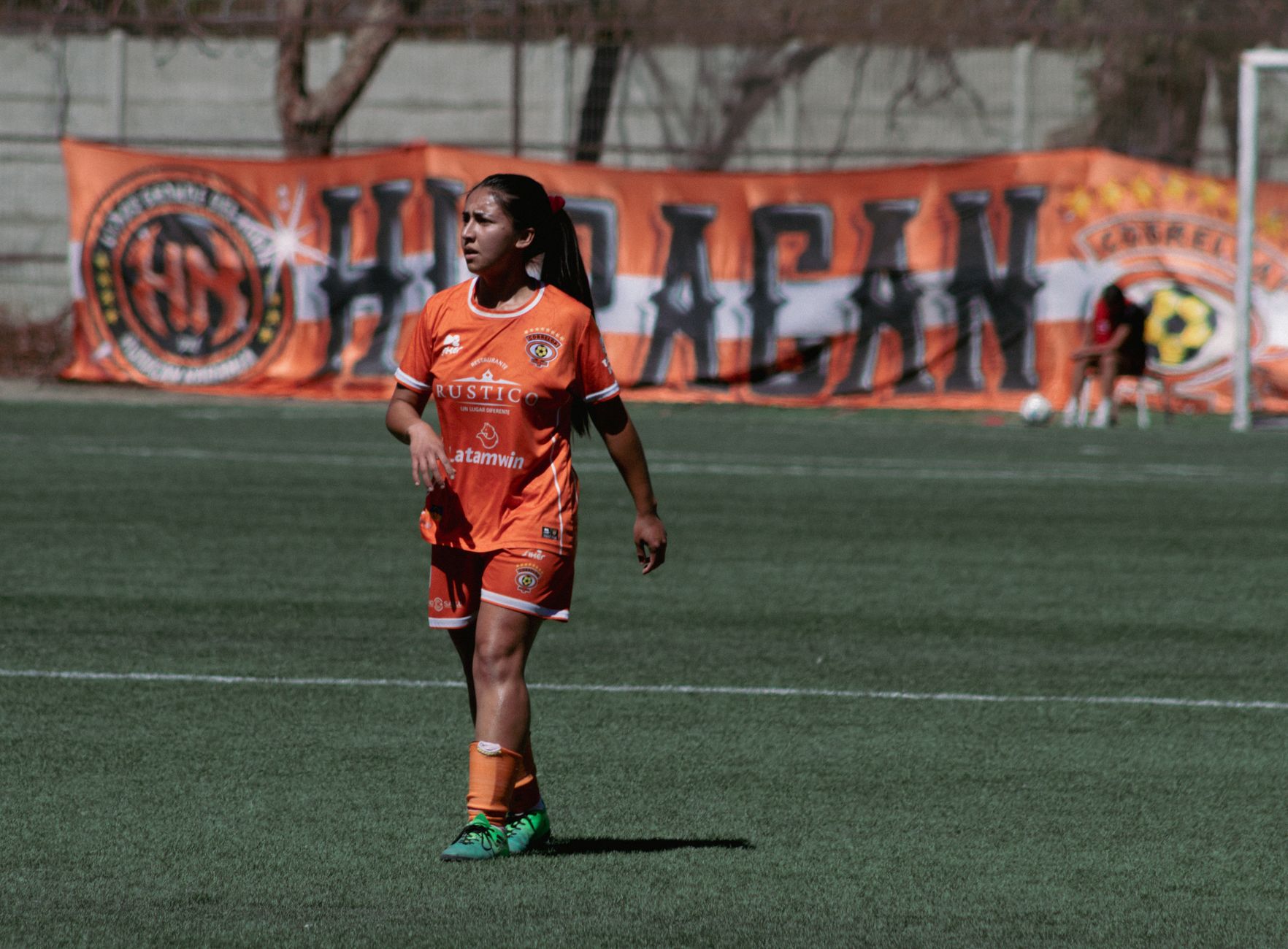 Female soccer player in orange uniform on outdoor field with banner background.