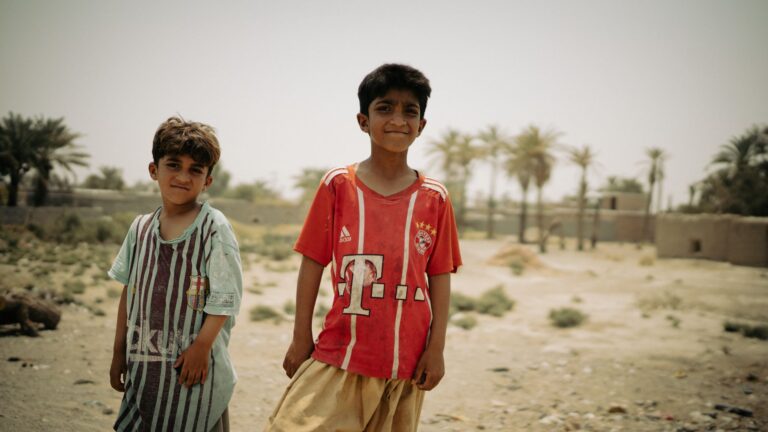 Two young boys enjoying a sunny day outdoors in Iranshahr, Iran with palm trees in the background.