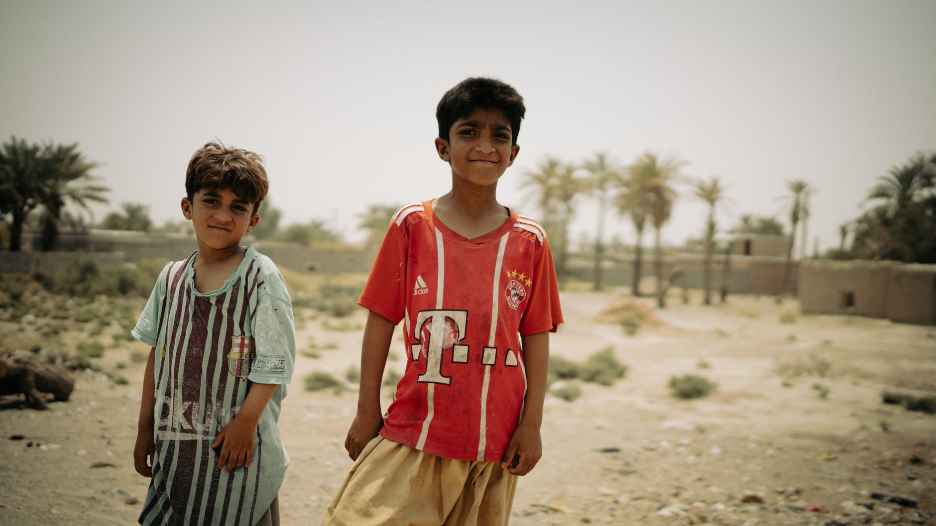 Two young boys enjoying a sunny day outdoors in Iranshahr, Iran with palm trees in the background.
