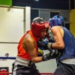 Two athletes in protective gear engage in a spirited boxing match inside a gym.