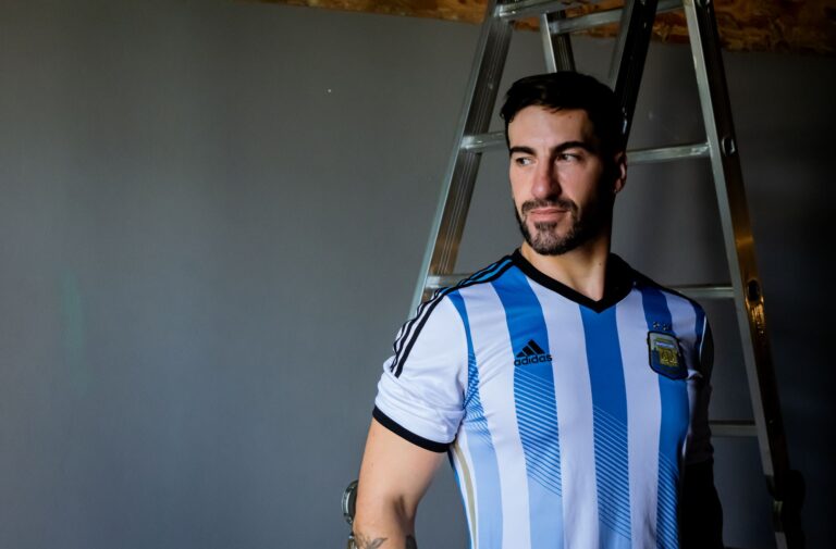 An adult male wearing an Argentina soccer jersey stands beside a ladder indoors.