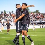 Two soccer players in black jerseys celebrate a goal on a sunny day at a packed stadium.