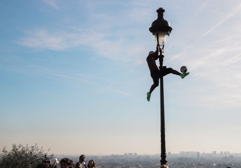 Silhouette of a man performing football tricks on a lamppost in Paris, with cityscape in the background.