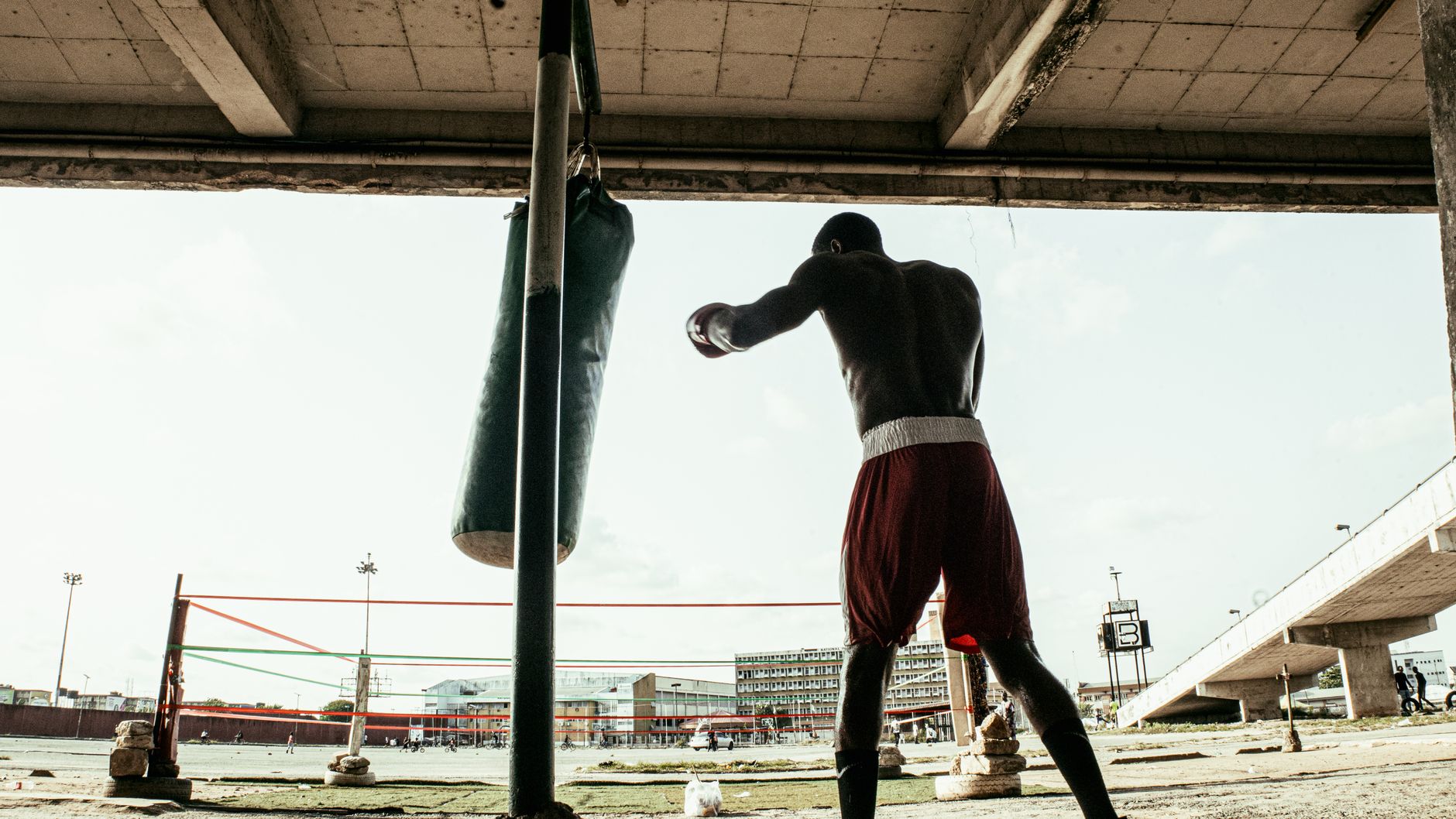 A boxer in red shorts practices punches with a hanging punching bag under a bridge in Ikeja, Nigeria.