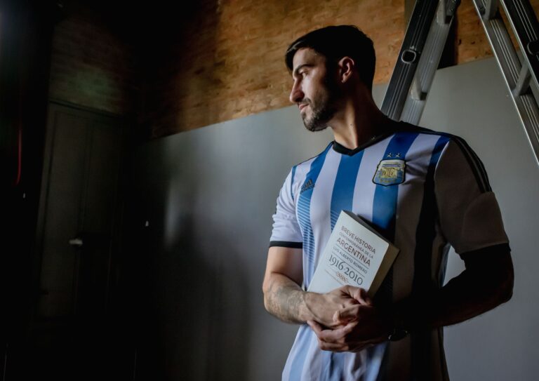 Man in Argentina soccer jersey holding book indoors, looking thoughtful.
