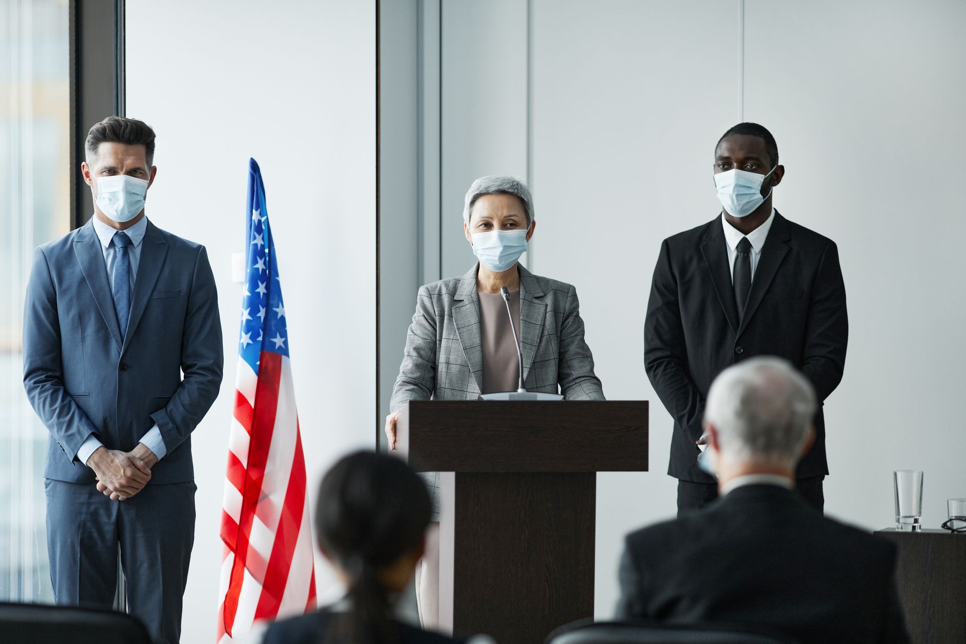 Masked business leaders at a conference podium with US flag backdrop, emphasizing safety and professionalism.