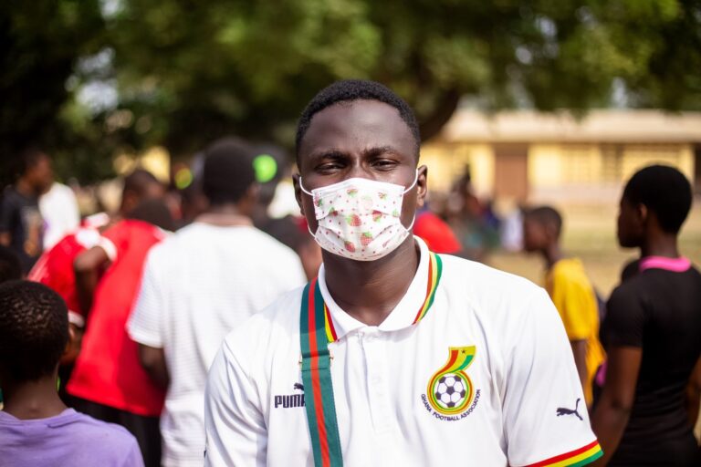 Young man wearing a mask at an outdoor event with a vibrant crowd in the background.