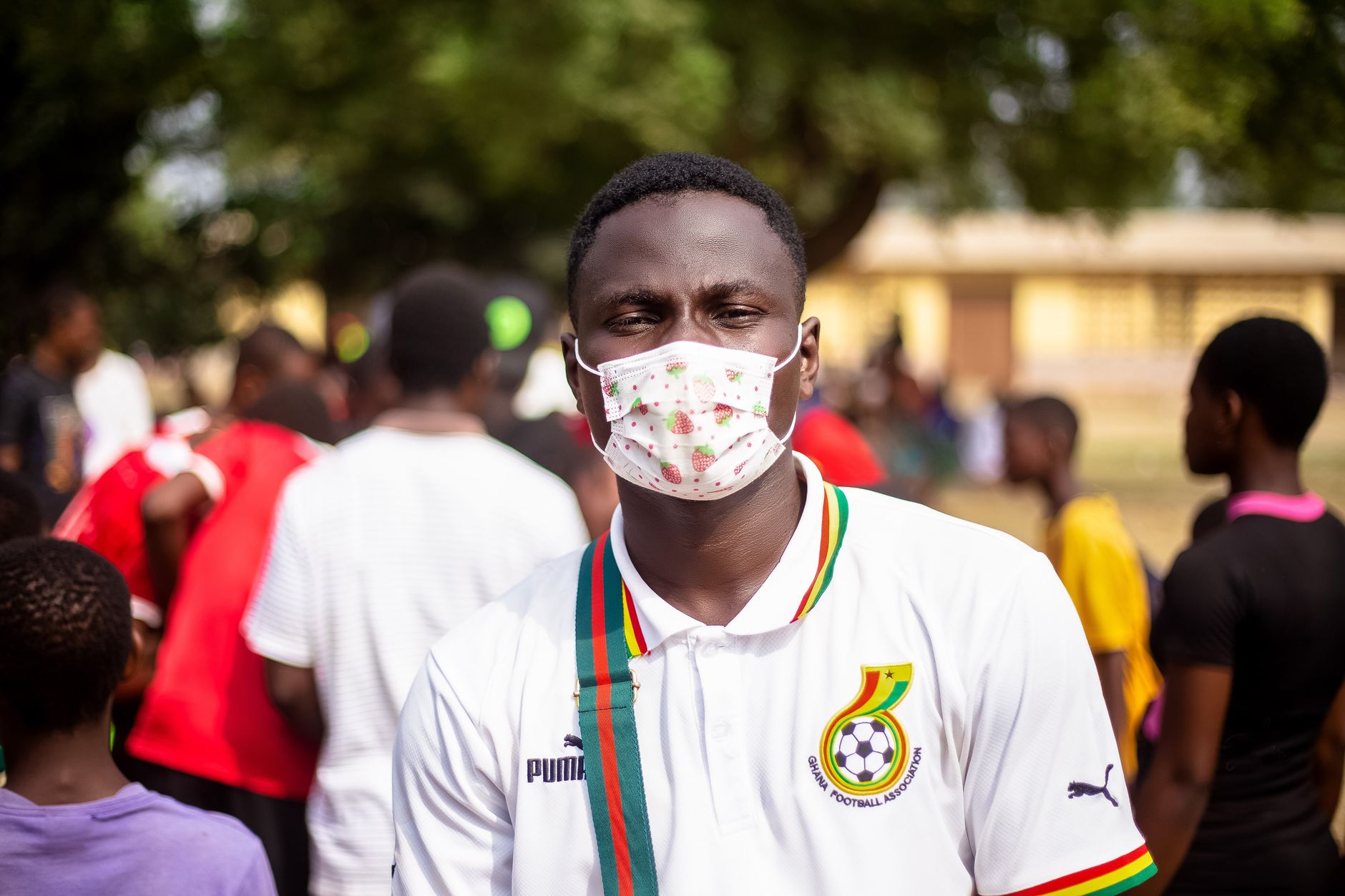 Young man wearing a mask at an outdoor event with a vibrant crowd in the background.
