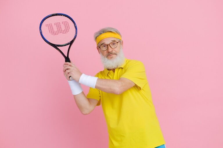 Elderly man in yellow sportswear holding a tennis racket against a vibrant pink background.