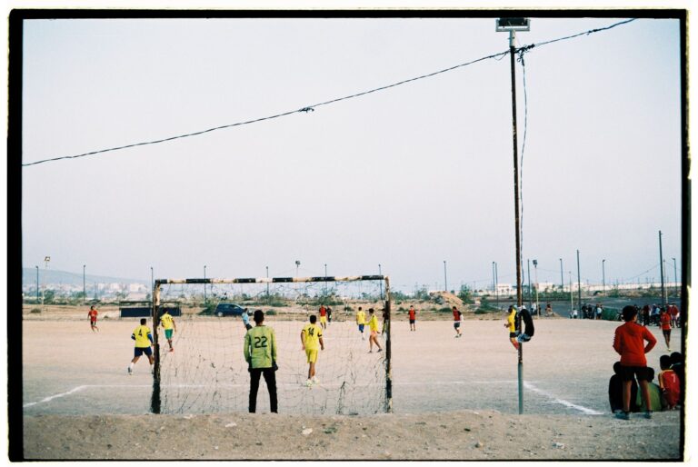 Players engaged in a soccer match on a dusty field in Morocco with scattered spectators.