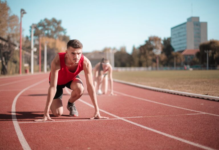 Concentrated male sprinter in soft focus standing on crouch start pose on track while blurred female athlete preparing for run on same position