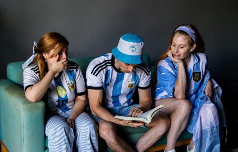 Three soccer fans in Argentina jerseys reading a book indoors