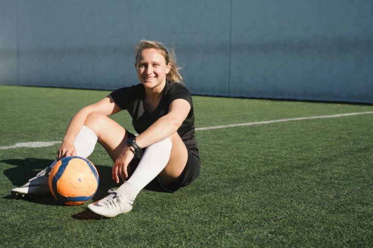 A smiling female soccer player sitting on a soccer field with a ball, enjoying a sunny day.