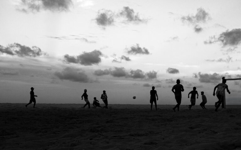 Silhouetted children play soccer on a sandy beach in Natal, Brazil during sunset.