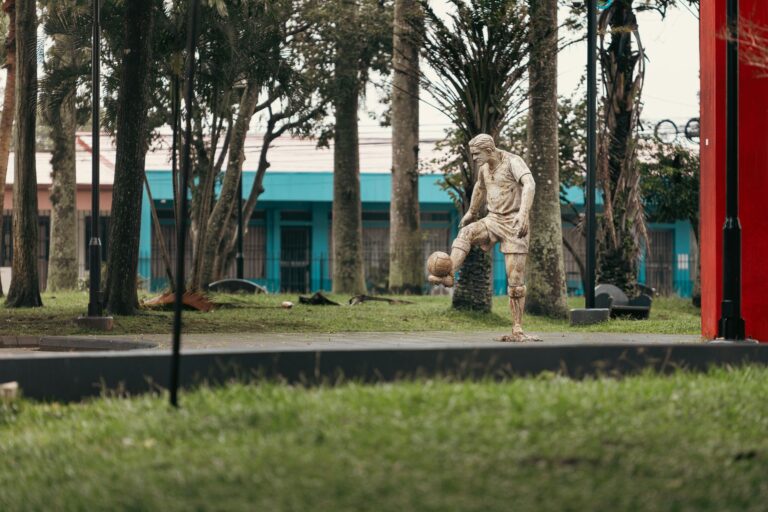 Stone sculpture of a soccer player juggling a ball in a lush Alajuela park setting.