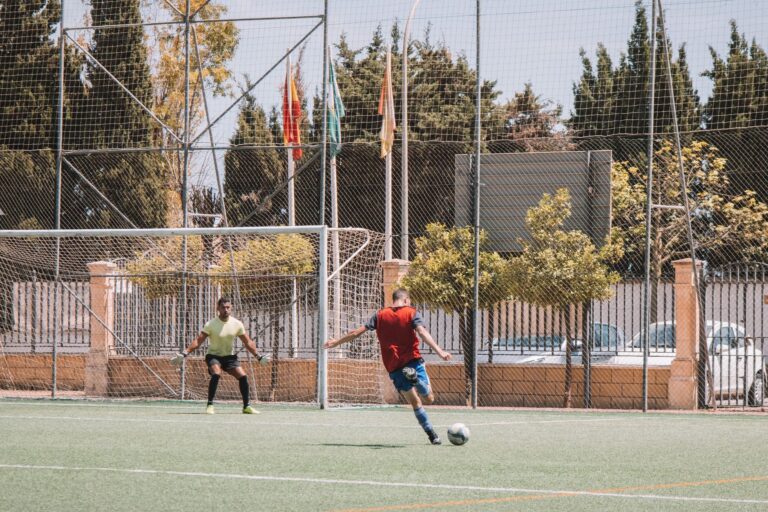 Dynamic soccer match in Málaga, Spain showing player kicking towards goal on sunny day.