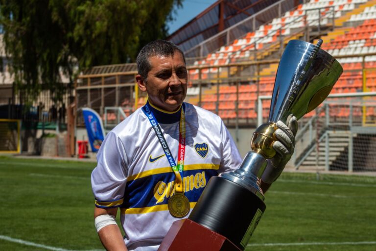 Adult soccer player holding a championship trophy on a sunny day at an outdoor stadium.