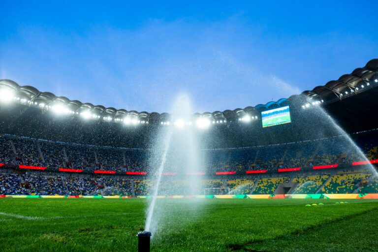 Nighttime view of a soccer stadium with sprinklers watering the field, enhancing the vibrant atmosphere.