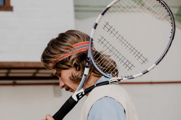 Tennis player with long hair wearing a sweatband holds racket on shoulder outdoors.