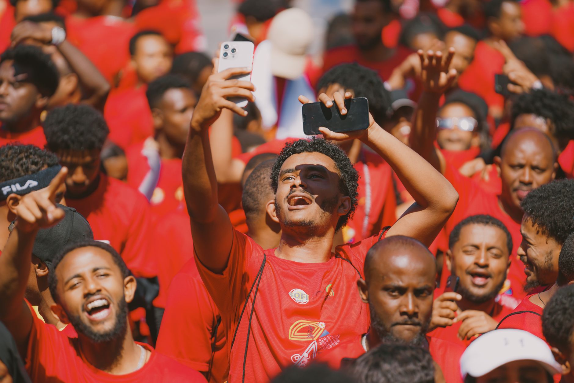 Joyful crowd of adults dressed in red holding smartphones, celebrating outdoors during the day.