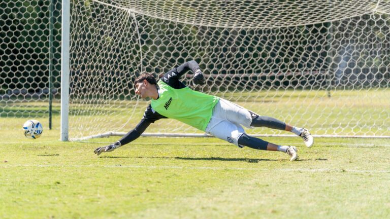 Athletic goalkeeper diving to stop a soccer ball during a sunny outdoor game.