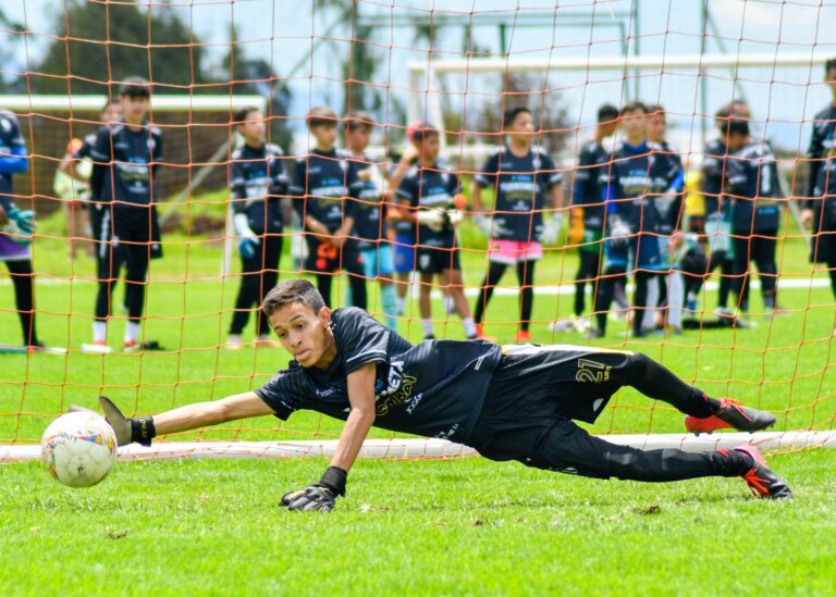 A young goalkeeper dives to save a soccer ball during a practice match outdoors.