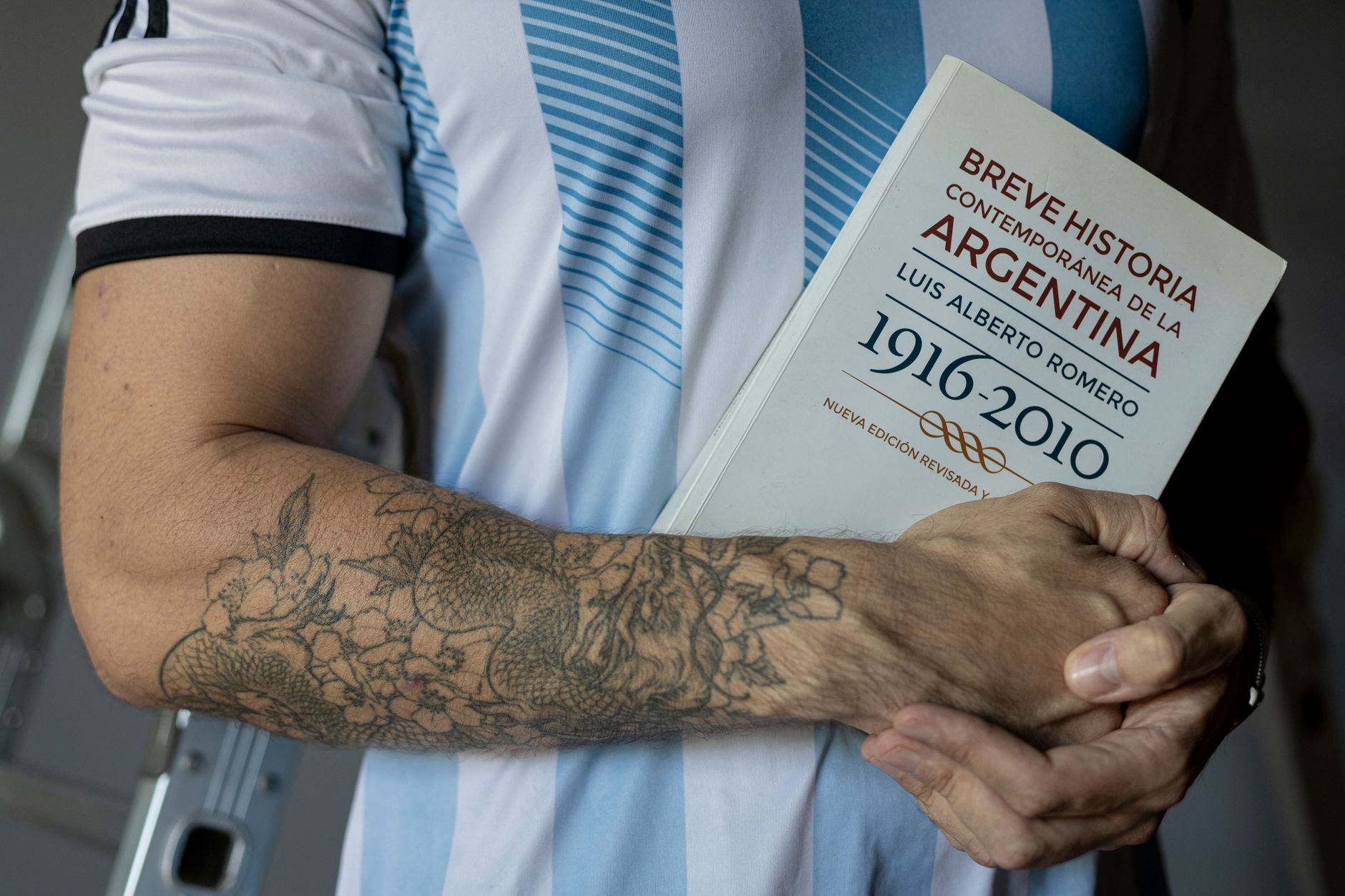 A tattooed arm holding a book about Argentina's history wearing a team jersey.
