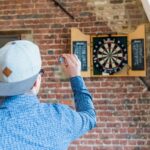 An adult man in a blue shirt playing darts indoors, aiming at a brick wall-mounted dartboard.