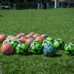 A collection of colorful soccer balls on a green field in Brasília, Brazil, under a clear blue sky.