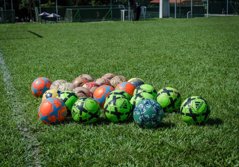 A collection of colorful soccer balls on a green field in Brasília, Brazil, under a clear blue sky.