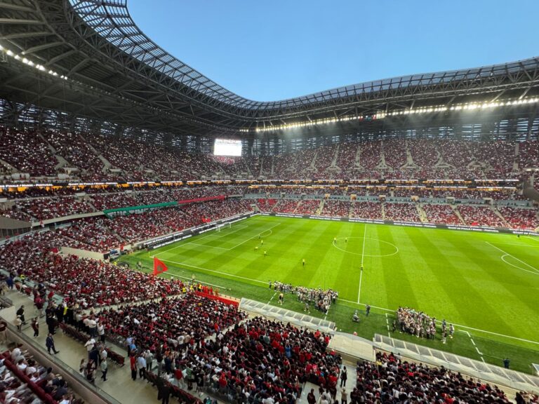 A stadium full of cheering fans watching a live football match during daylight.