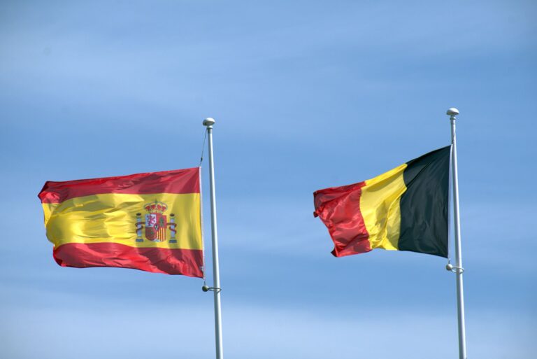 Flags of Spain and Belgium waving on poles under a blue sky, symbolizing international relations.