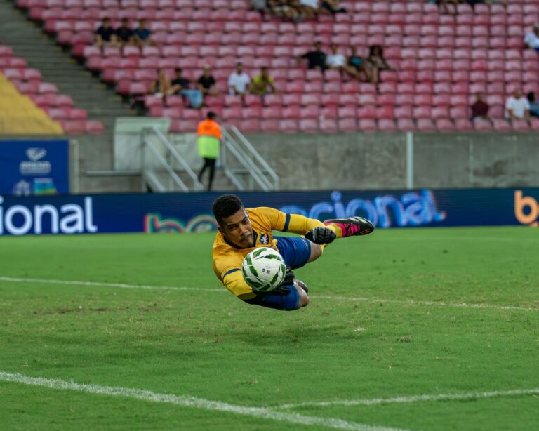 A goalkeeper in action diving to catch a soccer ball during a match in a large stadium.