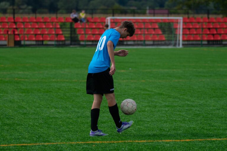Teenage soccer player skillfully balancing a soccer ball on a grassy field with red stands.