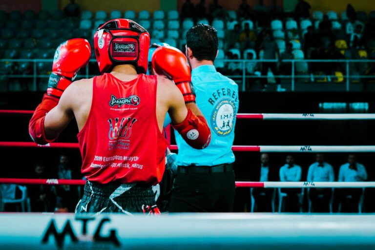 Rear view of a Muay Thai boxer in red gear facing the referee inside a boxing ring.