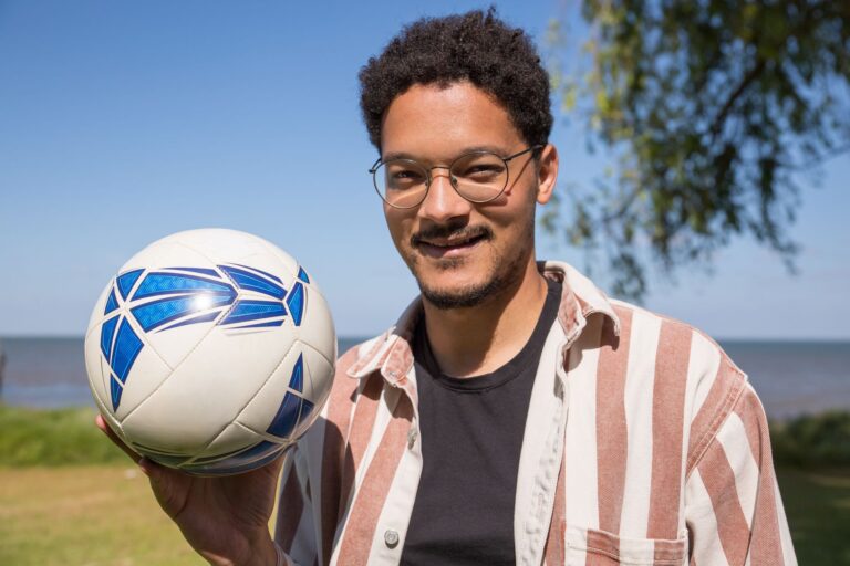 A smiling man with glasses and striped shirt holding a soccer ball outdoors in Portugal.