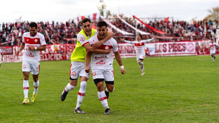 Soccer players celebrating a goal on an outdoor field with cheering crowd.