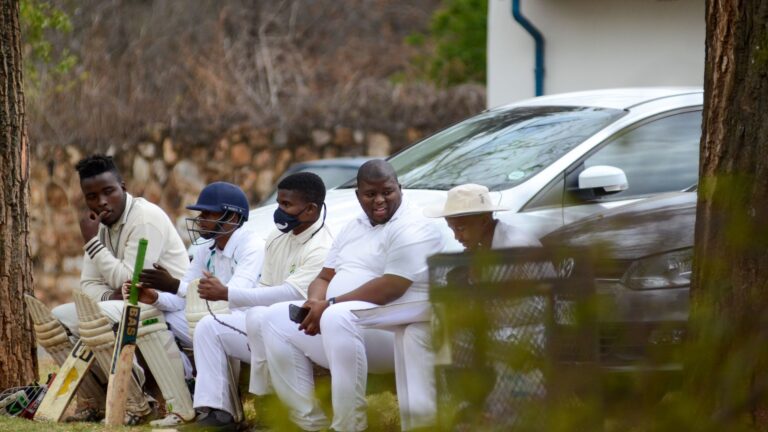 Group of cricket players sitting on a bench outdoors, dressed in white uniforms.