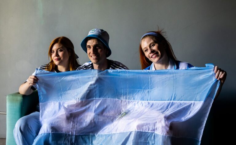 Three people holding the Argentine flag indoors. Celebratory and patriotic mood.