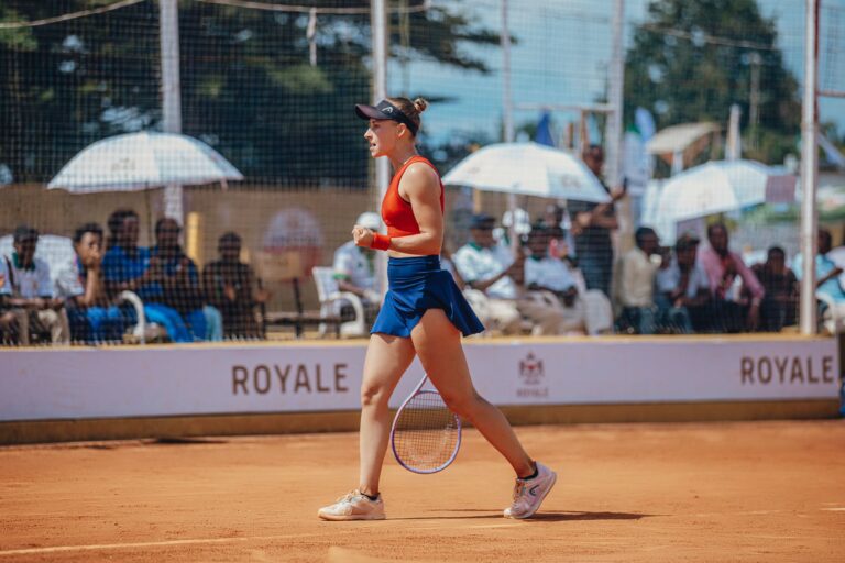 A triumphant female tennis player in action on a clay court with spectators in the background.