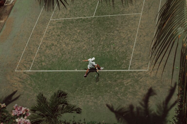 Aerial shot of a tennis player mid-action on a tropical court surrounded by lush vegetation.
